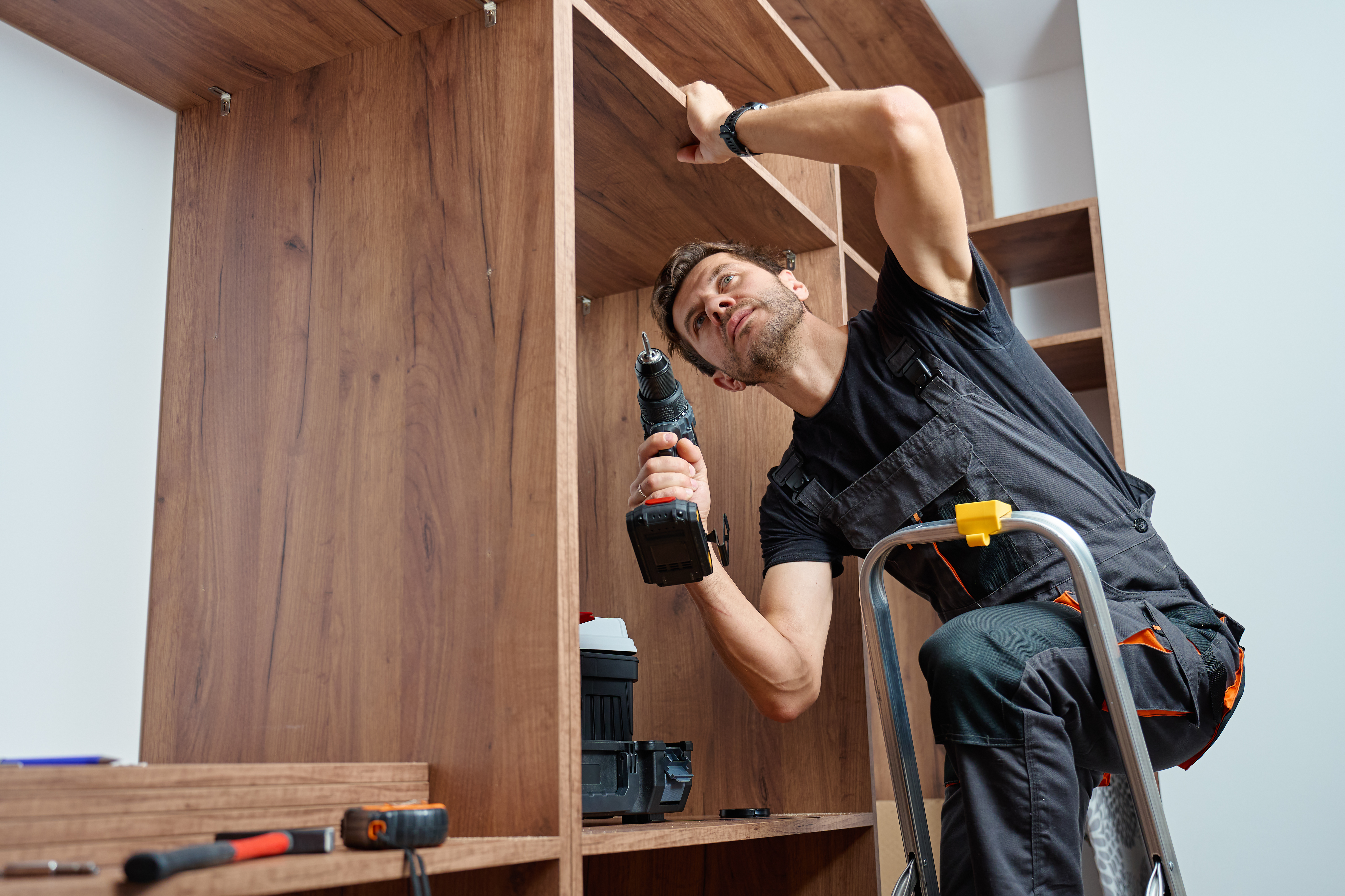 Man wearing work overalls assembling wooden wardrobe with drill and tools. Carpenter standing on step ladder, furniture installation process